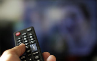 A hand holding a remote control presses buttons to switch TV channels