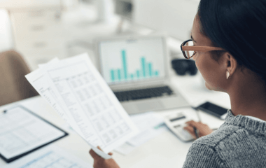 A woman looking through pages of data while working on her computer.