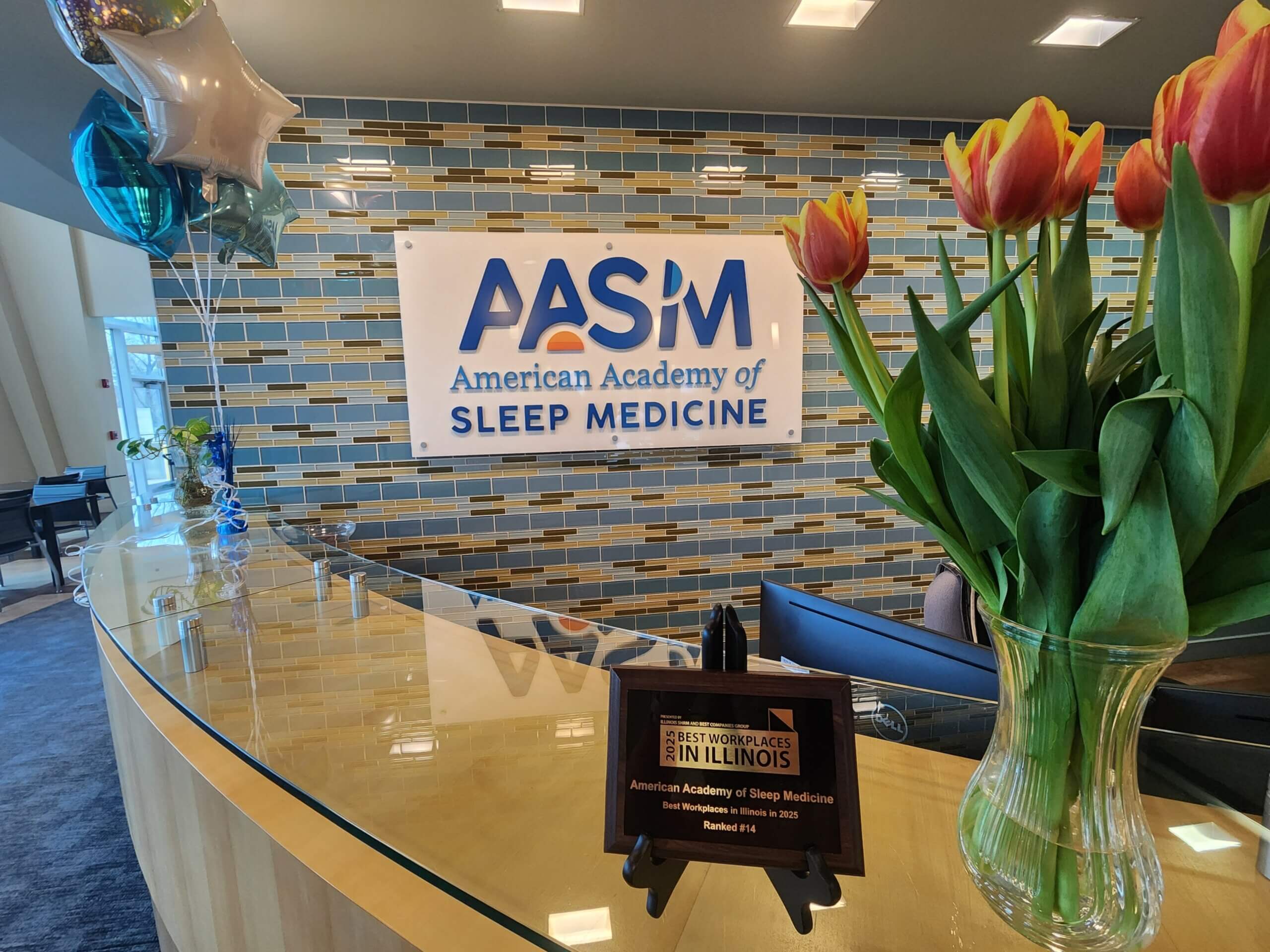 A display featuring a sign for the American Academy of Sleep Medicine (AASM) on a tiled wall, accompanied by a colorful balloon arrangement. The balloons include a star-shaped design and a speech bubble with the message, 'Thanks! (for all you do).'
