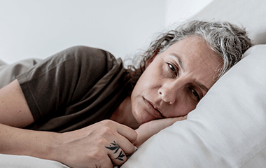 Person lying on their side in bed with eyes closed, resting on a pillow, suggesting sleep or fatigue.