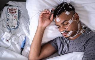 Person lying in bed wearing sleep monitoring sensors and wires connected to medical equipment, indicating an overnight sleep study or sleep disorder evaluation.